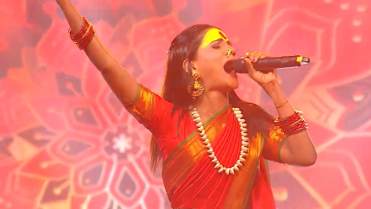 Barnali Hota singing on stage in a red saree, arm raised